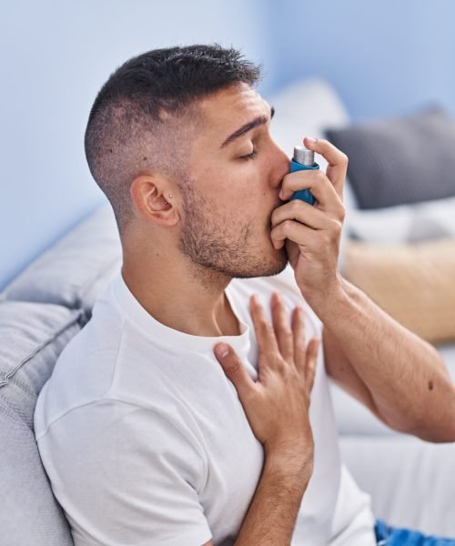 Young hispanic man using inhaler sitting on sofa at home